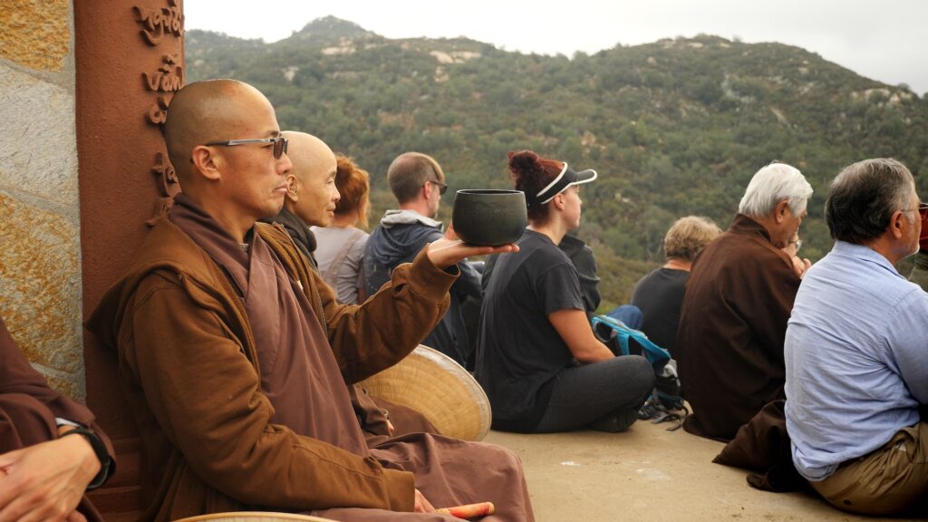 Monastic with bell and mountains in the background. Lay friends sitting all around at the stupa of Deer Park Monastery.